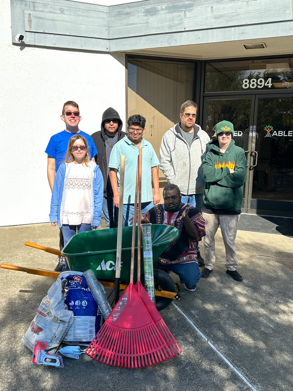 ABLED clients posing in front of ABLED's headquarters, in front of gardening and landscaping tools (wheelbarrow, dirt, rakes)